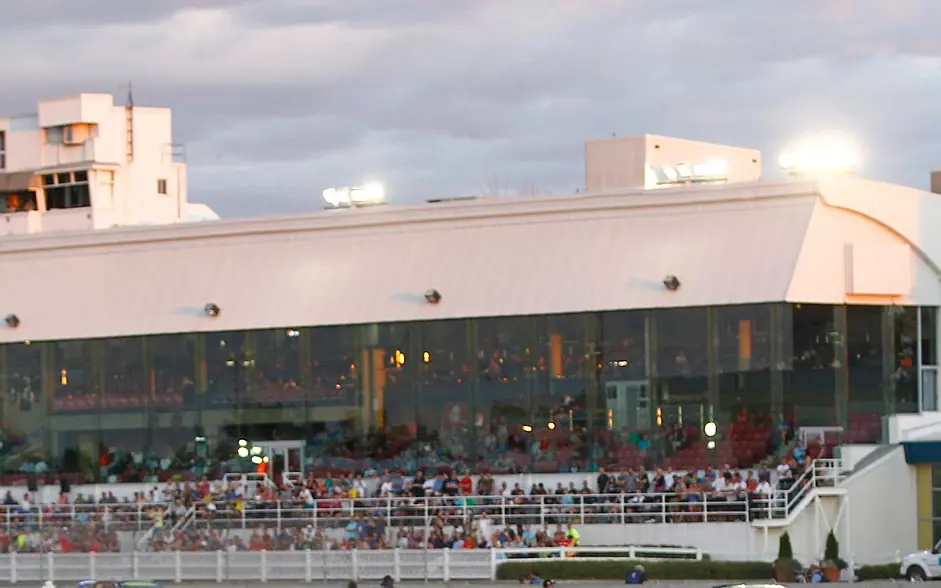 The glass-fronted grandstand of the Hippodrome de Trois-Rivières with spectators along the rail, at dusk